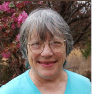 An older woman with gray hair and glasses smiles at the camera. Member of the Month Stefani Beason is wearing a light blue shirt and stands outdoors in front of a blooming pink flower bush. -WorkIT Coworking Center