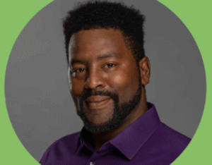 Marshon Carthen, Member of the Month, is a man with short, curly black hair and a trimmed beard, wearing a purple collared shirt and smiling slightly. The background is gray and circular with a green border. -WorkIT Coworking Center