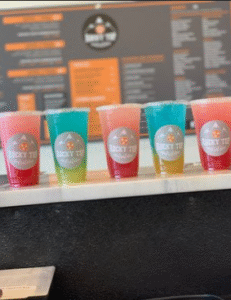 Five colorful drinks in clear plastic cups with Rocky Top Nutrition logos sit on a counter, arranged from left to right in yellow, red, pink, blue, and orange. A blurry menu hangs on the wall behind them at this favorite local florist near Stilly Floral. -WorkIT Coworking Center