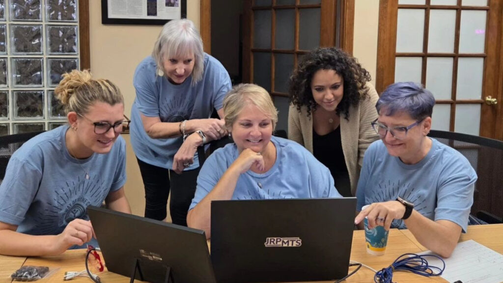 Five women in light blue shirts gather around a desk, smiling and looking at a laptop screen. Two are sitting and three are standing behind, appearing engaged and collaborative as they explore volunteer opportunities with United Way in a bright office setting. -WorkIT Coworking Center
