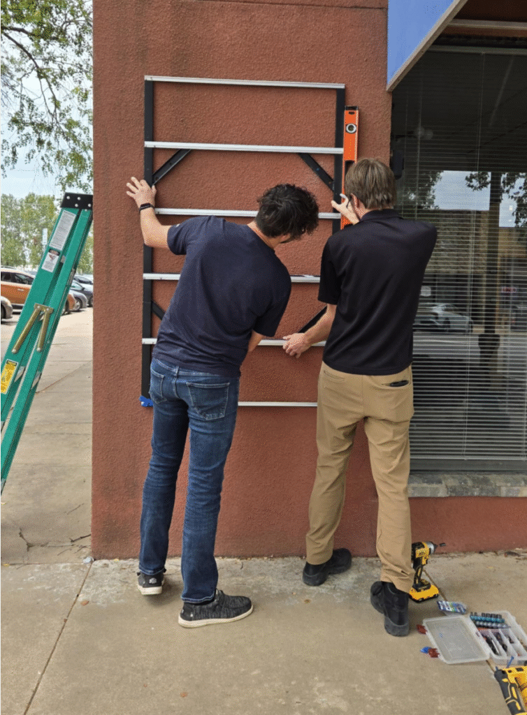 Last month, two men install a metal frame on an exterior wall, using a level tool for alignment. A ladder, power drill, and toolbox are nearby as they work beside a large window on the sidewalk. -WorkIT Coworking Center