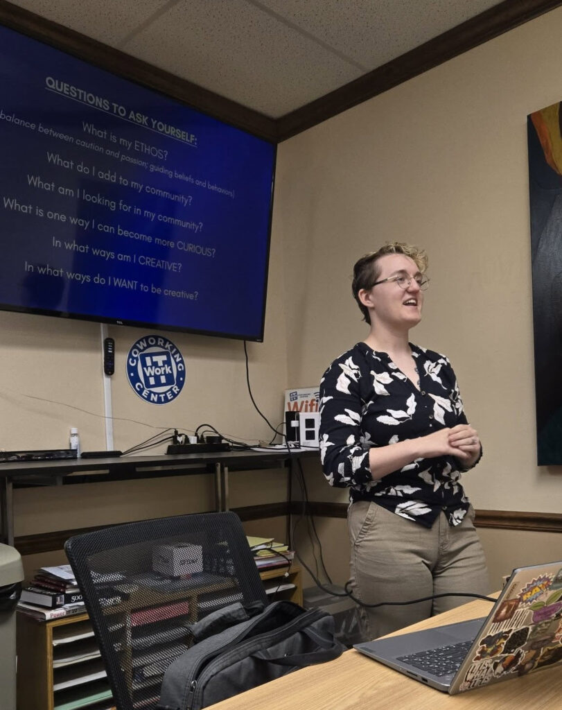 A person wearing glasses and a patterned shirt stands and smiles while presenting at a Lunch & Learn in a meeting room. Behind them, a screen displays reflective questions to ask yourself. A laptop and office supplies are visible on the table. -WorkIT Coworking Center