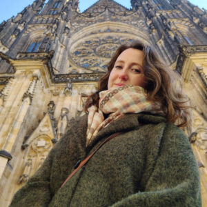 Member of the Month Marshon Carthen stands in a green coat and plaid scarf before an ornate Gothic cathedral, its detailed stonework and large stained glass window framing her gaze at the camera. -WorkIT Coworking Center