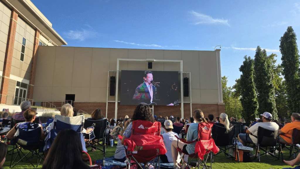 A crowd sits on lawn chairs facing an outdoor screen showing a man speaking at the September After Hours, with a large building and trees in the background under a clear blue sky. -WorkIT Coworking Center