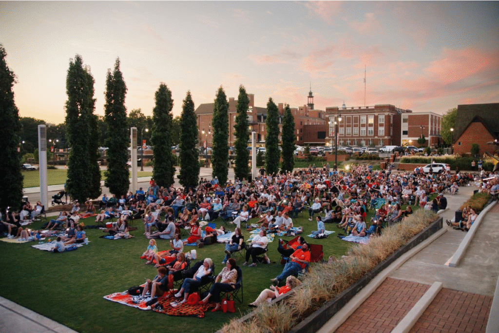 A large crowd of people sits on blankets on a grassy lawn at sunset, gathered for September After Hours—an outdoor event surrounded by tall trees and brick buildings in the background, featured in WorkIT News. -WorkIT Coworking Center