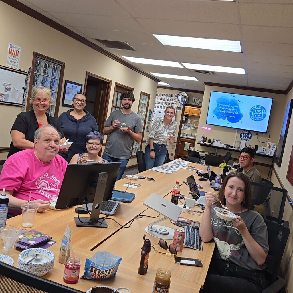A group of eight people smiling and enjoying food around a conference table during a Lunch & Learn in the office, with laptops, snacks, drinks, and a presentation screen visible in the background. -WorkIT Coworking Center
