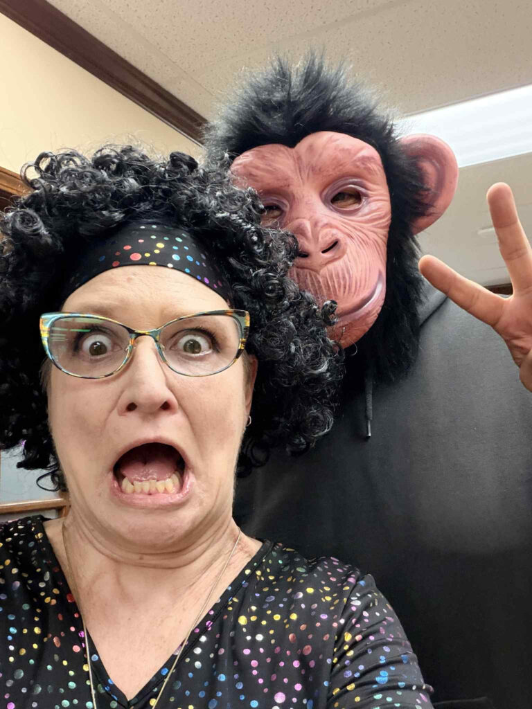 A woman with curly hair, glasses, and a surprised expression stands in front of a person wearing a monkey mask and making a peace sign at last month’s Lunch & Learn. Both appear to be dressed in costumes. -WorkIT Coworking Center