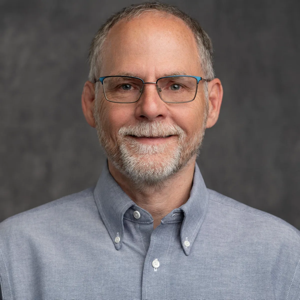 A middle-aged man with short gray hair, a beard, and glasses, wearing a light blue button-up shirt, smiles in front of a plain gray background as he is recognized as Member of the Month by Beth Chaffin. -WorkIT Coworking Center