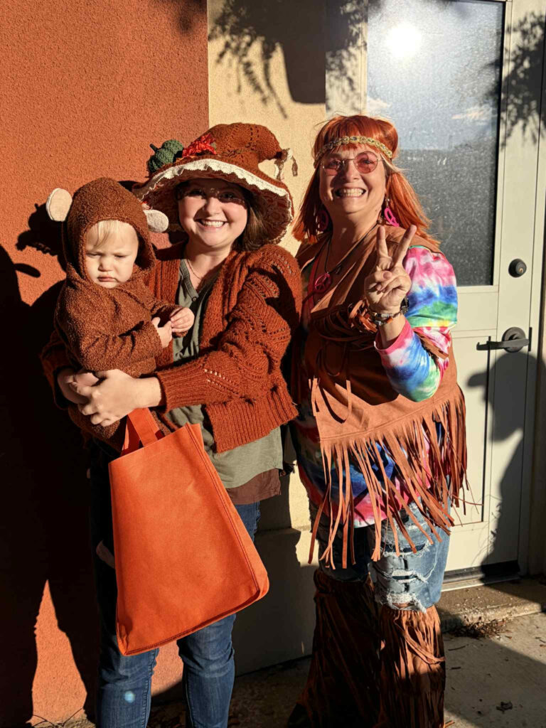 Three people in costume stand outside after a Lunch & Learn. One adult holds a baby dressed as a monkey, both in brown outfits. The other wears a colorful tie-dye shirt, fringe vest, sunglasses, and flashes a peace sign in the bright sunlight. -WorkIT Coworking Center