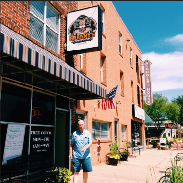 An older man stands outside Granny’s Kitchen & Catering, a brick restaurant reminiscent of Louie’s Grill & Bar, with an American flag, open sign, and benches along the sidewalk on a sunny day. -WorkIT Coworking Center