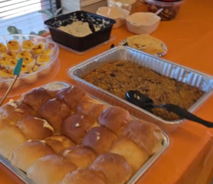A table covered with an orange cloth holds a tray of dinner rolls, a casserole, deviled eggs, dips, chips, and other dishes—perfect for a Friendsgiving 2025 meal or gathering featured by WorkIT News. -WorkIT Coworking Center