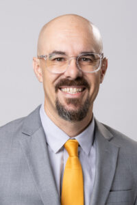 Shane Berry, our Member of the Month, is a bald man with a goatee and mustache, wearing clear glasses, a light gray suit, a light blue shirt, and a yellow tie, smiling at the camera against a plain light background. -WorkIT Coworking Center