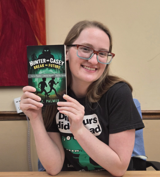 A smiling person with glasses holds up a book titled Hunter and Casey Break the Future by D. Palmer. They wear a black T-shirt that says Dinosaurs did not read. A colorful fabric hangs on the wall behind them at WorkIT News. -WorkIT Coworking Center