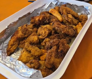 A white tray lined with aluminum foil holds several pieces of golden-brown fried chicken, perfect for sharing at your Friendsgiving 2025 celebration, placed on a wooden surface. -WorkIT Coworking Center