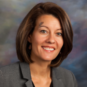 Jamie Andrews, our Member of the Month, smiles warmly in front of a blurred, multicolored background. She has straight brown hair and wears a gray blazer with elegant earrings. -WorkIT Coworking Center