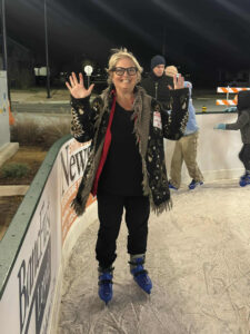 A smiling woman with glasses and a patterned coat stands on an outdoor ice skating rink at night, holding her hands up during a WorkIT News Holiday Party. Other skaters and street signs are visible in the background. -WorkIT Coworking Center