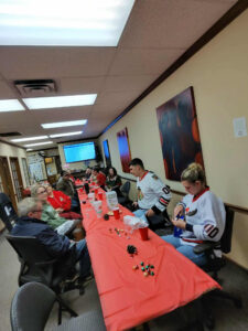 A group of people sits around a long table covered with a red tablecloth in an office setting for a Holiday Party. Some wear hockey jerseys, drinks and small items are scattered on the table, and two large abstract paintings hang on the wall. -WorkIT Coworking Center