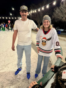 Two people ice skating outdoors at night, smiling at a holiday party by the rink’s edge. The man wears a white t-shirt, light jeans, and cap; the woman dons a festive moose hockey jersey, gray hat, and light jeans under twinkling string lights. -WorkIT Coworking Center