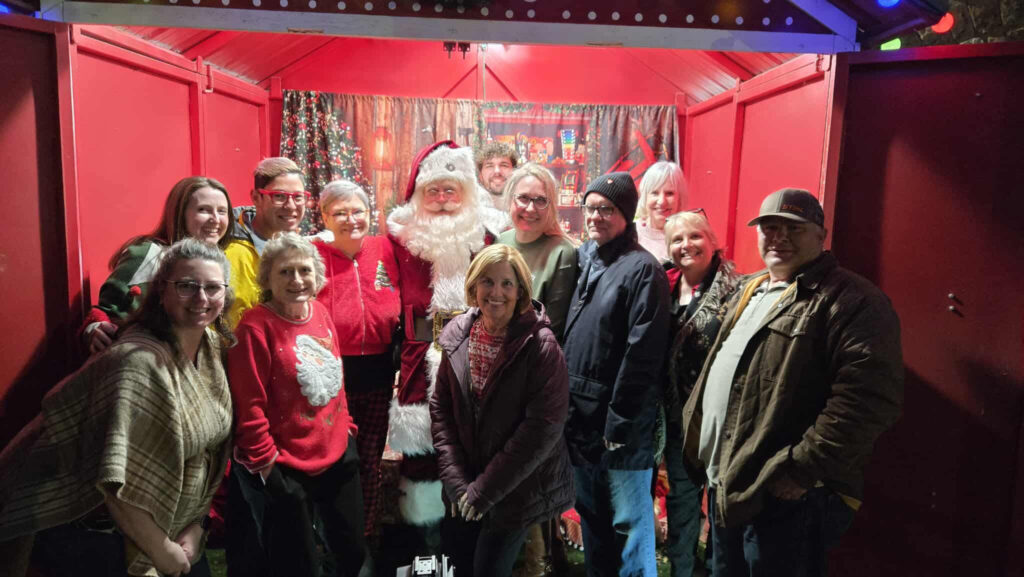 A group of people, both young and old, gather around Santa Claus inside a festive, red holiday booth at the WorkIT News Holiday Party, decorated with Christmas lights and ornaments. Everyone is smiling and dressed warmly for winter. -WorkIT Coworking Center