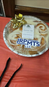 A round foil container of cinnamon rolls with icing sits on a red tablecloth, perfect for any Holiday Party. A gold bow decorates the lid with a business card, a JRPMTS LLC sticker, and black plastic tongs nearby. -WorkIT Coworking Center