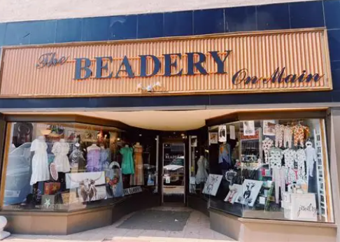 Storefront of The Beadery On Main, featuring large display windows with clothing, accessories, and various decor items arranged inside. The elegant script sign above the entrance reflects a touch of wealth and style on Main Street. -WorkIT Coworking Center