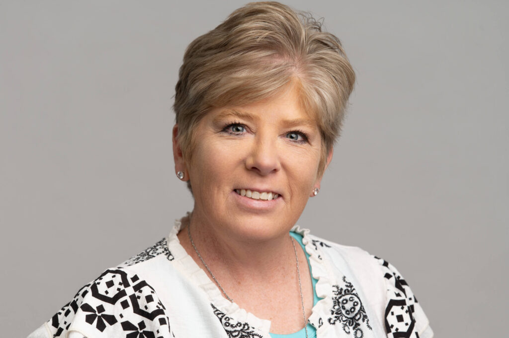 Member of the Month Keri Garrison smiles at the camera, her short light brown hair framing her face. She wears a white top with black patterns and small stud earrings, set against a neutral gray background. -WorkIT Coworking Center