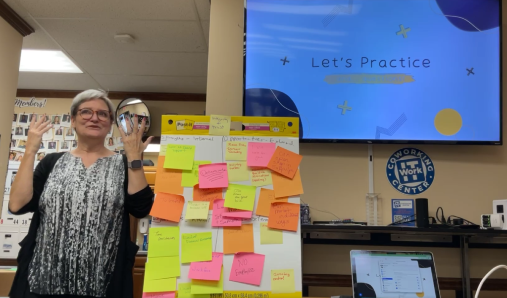 A woman stands and gestures beside a whiteboard covered in colorful sticky notes. Behind her, a large screen displays "Lets Practice" in a bright font—setting the scene for the September Lunch & Learn featured in WorkIT News. -WorkIT Coworking Center