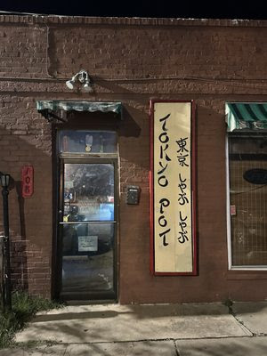 A brick building at night features a Tokyo Pot sign in English and Japanese. A glass door sits on the left, while a green awning covers a window, inviting any Neighbor-in-the-Know to this cozy Tokyo restaurant. -WorkIT Coworking Center