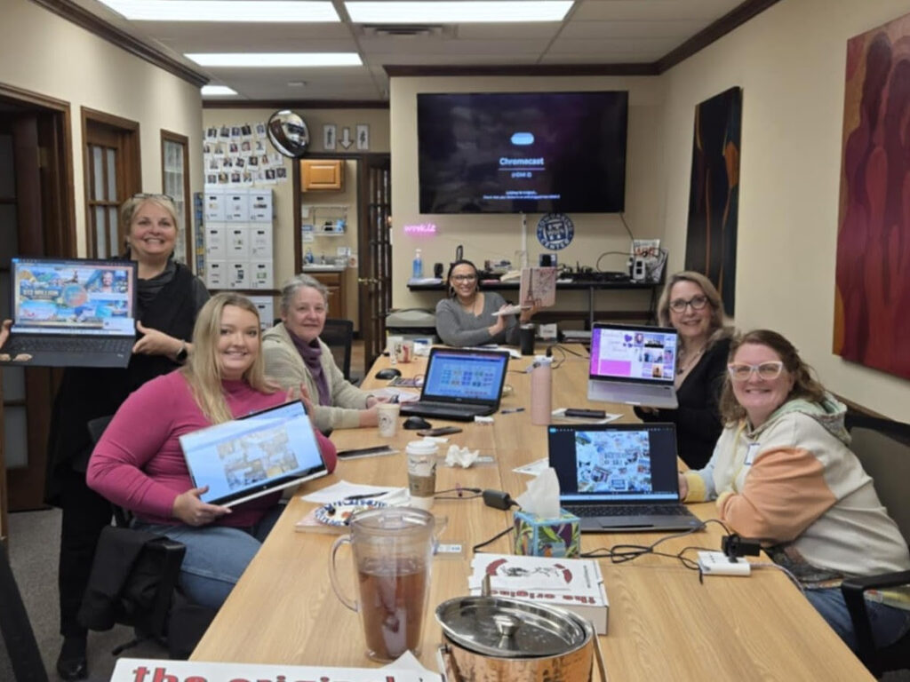 Six women sit around a conference table, each holding up a laptop displaying colorful screens from a digital vision board workshop. They are smiling in a cozy office space with drinks, papers, and WorkIT News featured on the TV in the background. -WorkIT Coworking Center