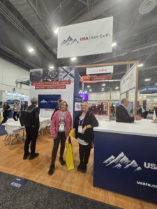Two women stand and smile at a trade show booth for USA Rare Earth, featured in Kristen's Corner, with banners reading “Securing Your Rare Earth Magnet Supply Chain.” Other attendees and booths are visible in the background. -WorkIT Coworking Center