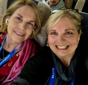 Two women smiling and sitting close together on a bus, both wearing blue lanyards and earbuds. The woman on the left wears a red top, and the woman on the right wears a dark top—capturing a candid Kristen's Corner moment. Other passengers are visible in the background. -WorkIT Coworking Center