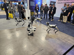 Three robotic dogs perform on a trade show floor as a group of people watch and take photos at Kristen's Corner. A large display screen and informational banners are visible in the background. -WorkIT Coworking Center