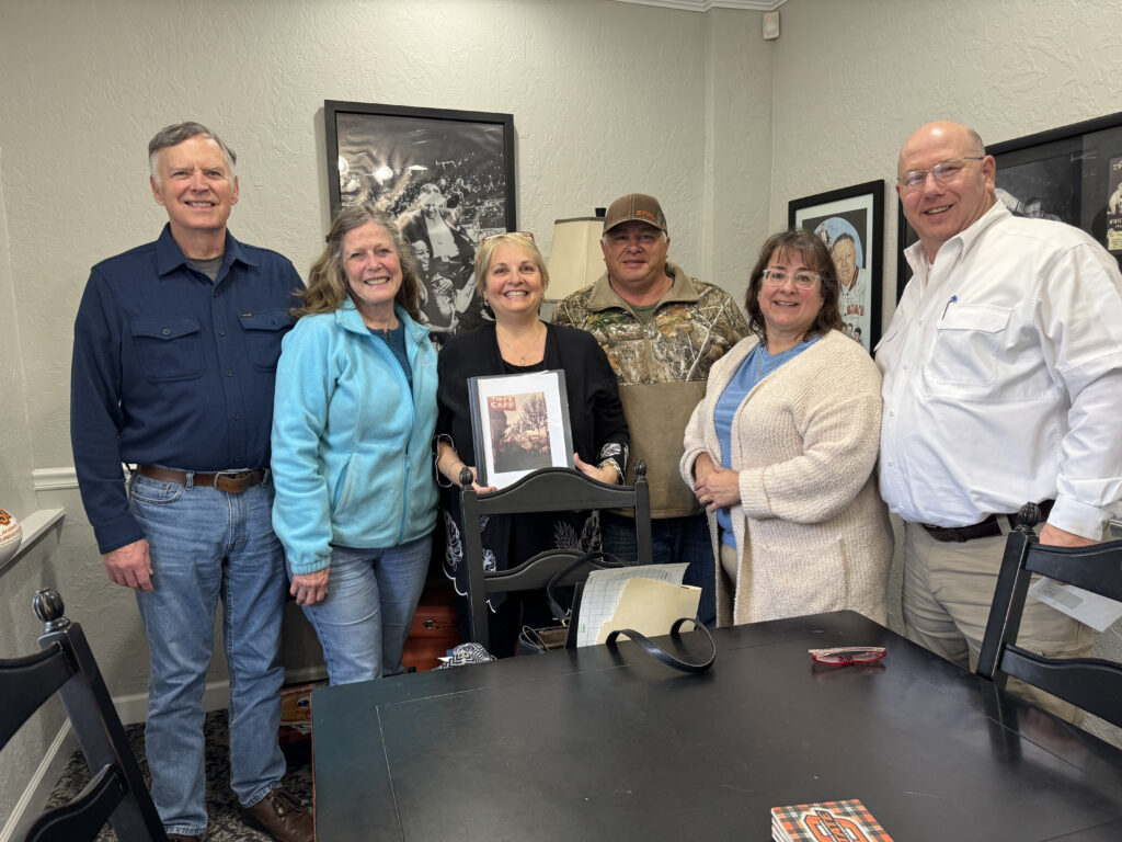 Six smiling adults stand together indoors around a table. The woman in the center holds a framed photo. Dressed in cozy, casual clothing, they gather for what looks like a Kristen's Korner meeting or friendly get-together. -WorkIT Coworking Center