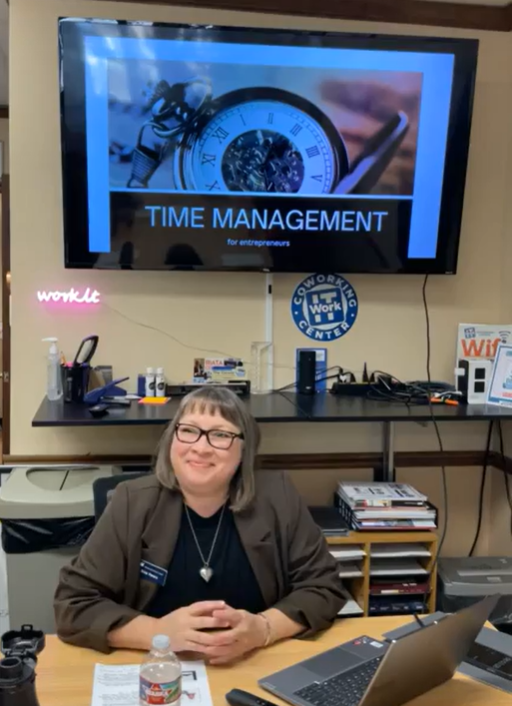 A woman sits at a desk, smiling, in front of a screen displaying a Time Management for Entrepreneurs presentation during the February Lunch & Learn. The desk holds books and drinks, with WorkIT News signs and equipment in the room. -WorkIT Coworking Center