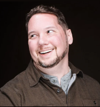 A man with short dark hair and a trimmed beard smiles while looking to his left. Member of the Month is noted as he wears a brown button-up shirt over a gray T-shirt against a dark background. -WorkIT Coworking Center