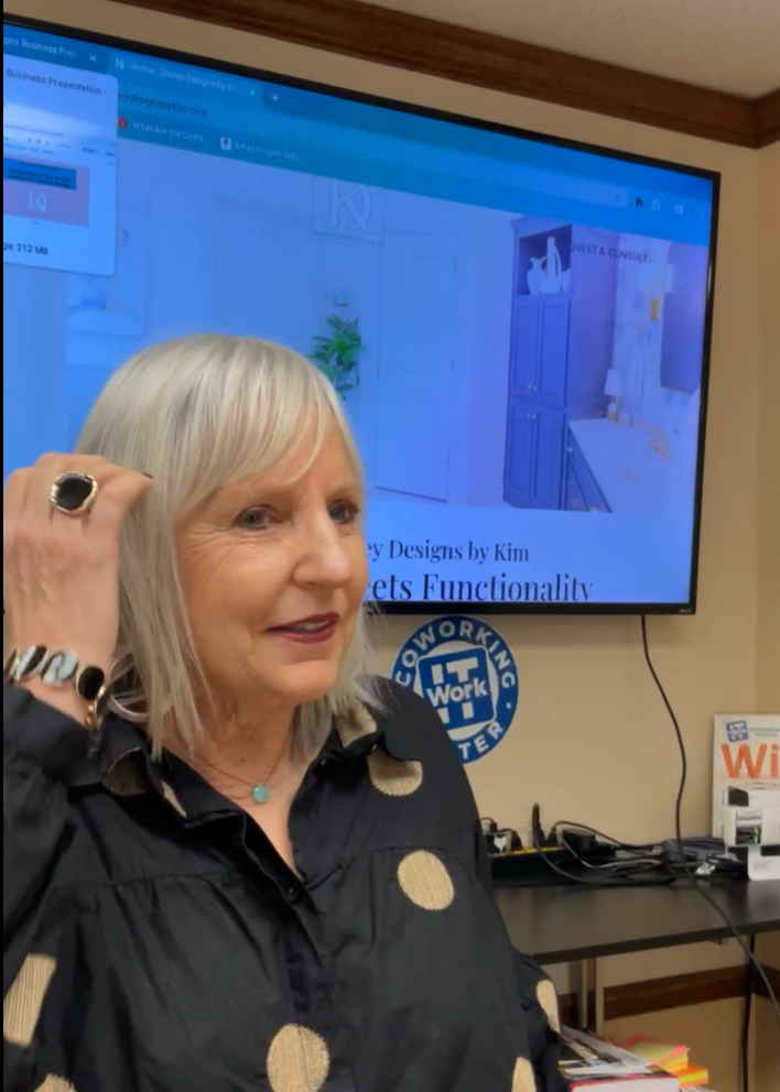 An older woman with light hair and a black polka-dot blouse stands before a large screen displaying a home décor website, likely presenting at the February Lunch & Learn. Books and plaques line the shelf behind her in this WorkIT News feature. -WorkIT Coworking Center