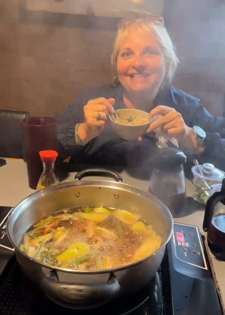 A smiling woman sits at a table holding a bowl at Kristen's Korner, with a steaming pot of vegetable and noodle soup on an induction stove in front of her. Various condiments and a glass of water are also on the table. -WorkIT Coworking Center