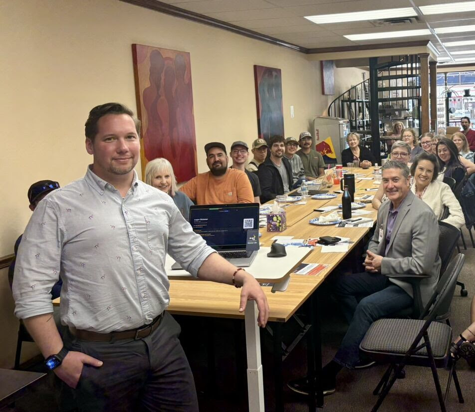 A group of people sit around a long conference table in an office, smiling at the camera during the March Lunch & Learn. One man stands at the end in the foreground. Papers, laptops, and drinks are on the table. Shelves and art decorate the room. -WorkIT Coworking Center