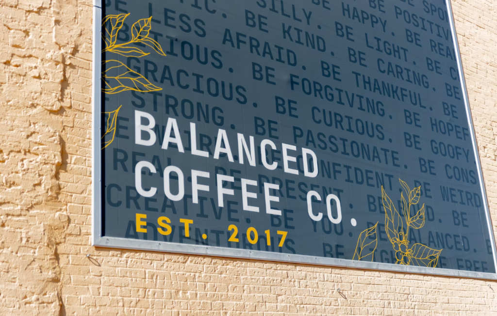 A large window sign for Balanced Coffee Co. Est. 2017, designed with yellow plant illustrations and motivational words, adds a welcoming foundation on a blue background to the tan brick wall—a real Neighbor-in-the-Know community touchpoint. -WorkIT Coworking Center
