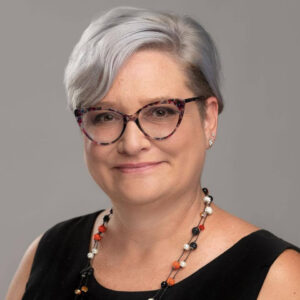 Member of the Month Rebecca Lowe, with short light gray hair and cat-eye glasses, smiles at the camera. She wears a sleeveless black top and a necklace with orange, black, and white beads against a plain gray background. -WorkIT Coworking Center