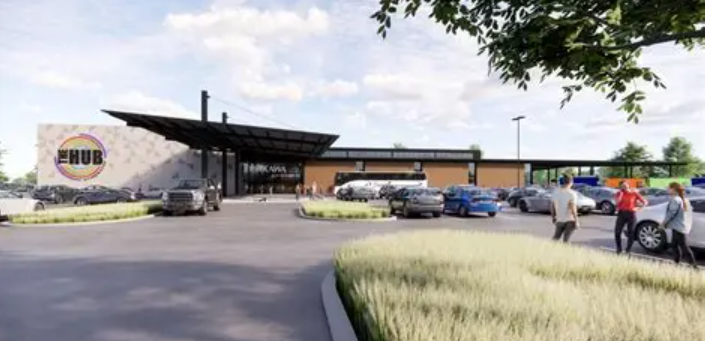 A modern community center labeled The Hub with large windows, a parking lot filled with cars, and people walking toward the entrance. A banner announces an upcoming Vision Board Workshop surrounded by trees and landscaped greenery. -WorkIT Coworking Center