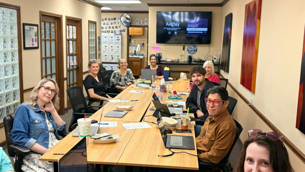 A group of people sits around a long conference table in an office during a WorkIT News Lunch & Learn in March, with laptops, papers, and drinks in front of them. A TV screen and filing cabinets are visible as everyone looks toward the camera. -WorkIT Coworking Center