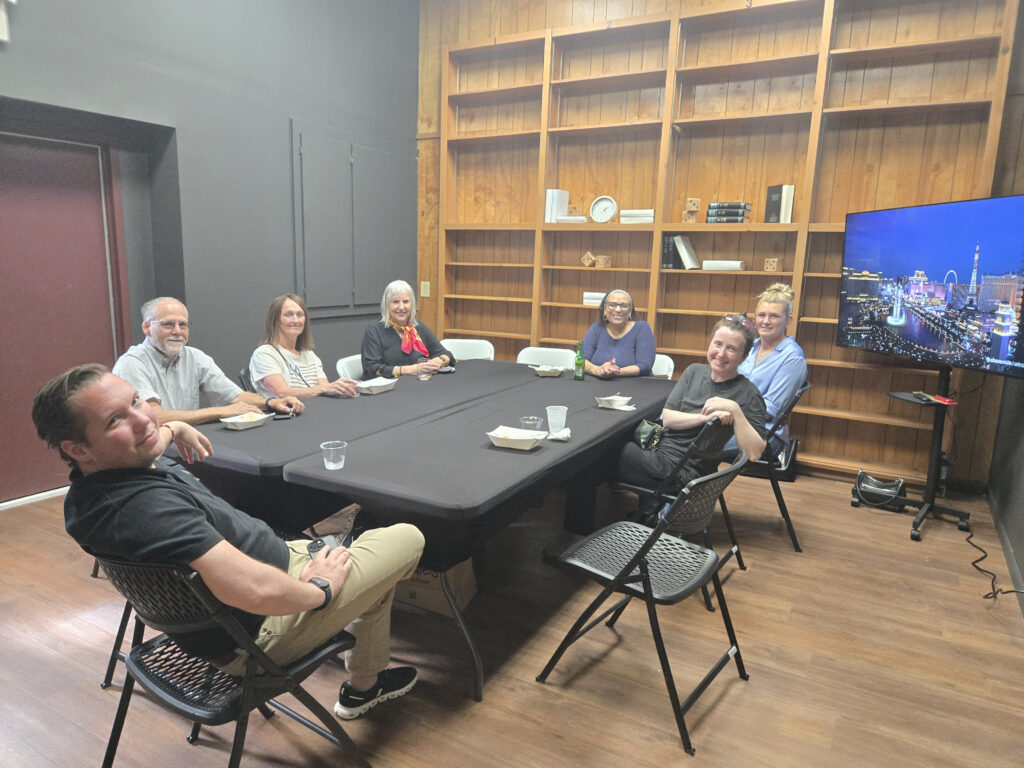 Seven people sit around a rectangular table in a room with wooden shelves and a TV displaying a cityscape. During the May Lunch & Learn, they appear to be having a meeting, with some food and drinks on the table. -WorkIT Coworking Center