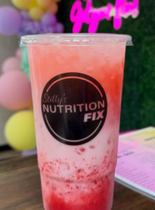 A pink and white iced drink in a clear cup labeled Stillys Nutrition Fix sits on a wooden table, with blurred pastel balloons and a neon sign reading “Neighbor-in-the-Know” glowing softly in the background. -WorkIT Coworking Center