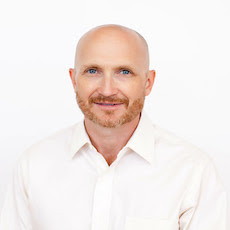 A middle-aged man with a bald head, light brown beard, and blue eyes, wearing a white collared shirt, poses against a plain white background as the proud Member of the Month. -WorkIT Coworking Center