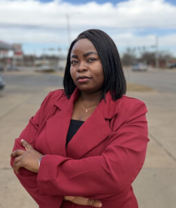 A woman wearing a red blazer stands outdoors with her arms crossed, looking confidently at the camera. The background shows a street, parked cars, and a cloudy sky, capturing the poised presence of Brian Hafner, Member of the Month. -WorkIT Coworking Center