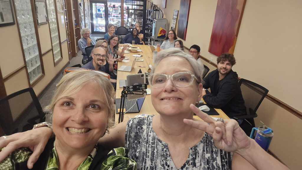 Two smiling women take a selfie in a conference room with a long table, surrounded by people sitting and facing the camera. One woman flashes a peace sign. Laptops and notebooks are on the table, capturing the collaborative spirit of May’s WorkIT News Lunch & Learn session. -WorkIT Coworking Center