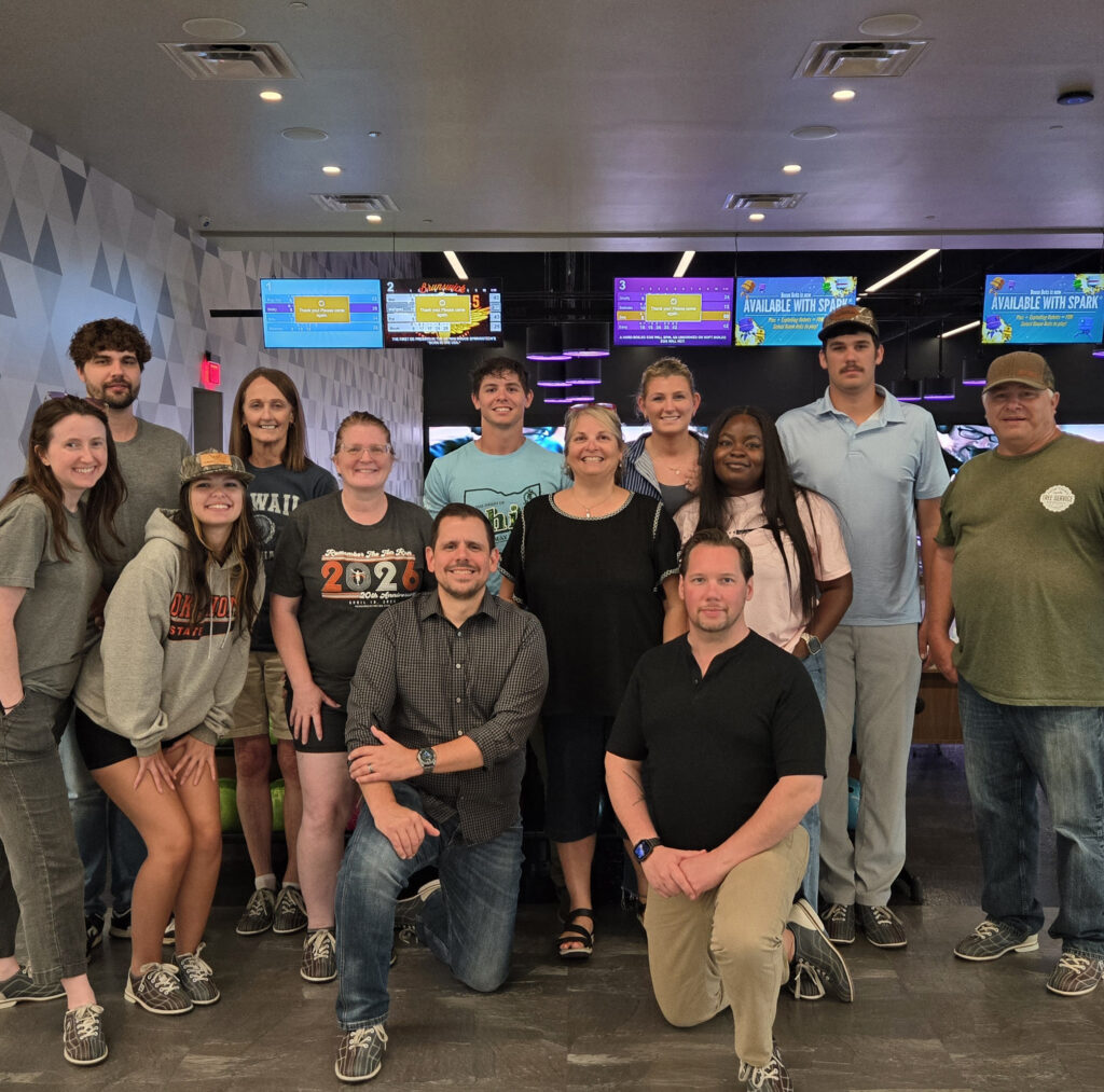 A group of thirteen people pose and smile together inside a modern bowling alley with colorful screens and bowling lanes in the background, celebrating a recent feature in WorkIT News. -WorkIT Coworking Center