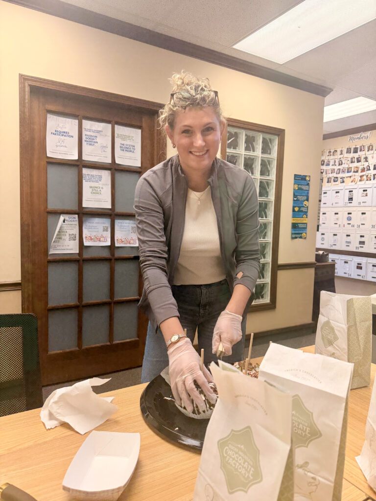 A person with curly hair, wearing gloves, a gray jacket, and a white shirt, smiles while cutting a cake at a table with bags and napkins in The HUB Stillwater office setting for WorkIT News. -WorkIT Coworking Center
