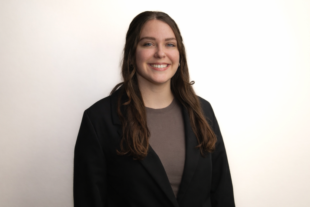 Caia Gorman, our Member of the Month, smiles warmly in front of a plain, light background, her long brown hair flowing over a black blazer and brown top. -WorkIT Coworking Center